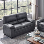 A modern living room setup featuring a dark gray faux-leather loveseat and a matching three-seater sofa, arranged around a glass-top coffee table on a blue-gray rug. The room has gray walls, a large multi-pane window looking out onto green trees, and a small console table with framed art.