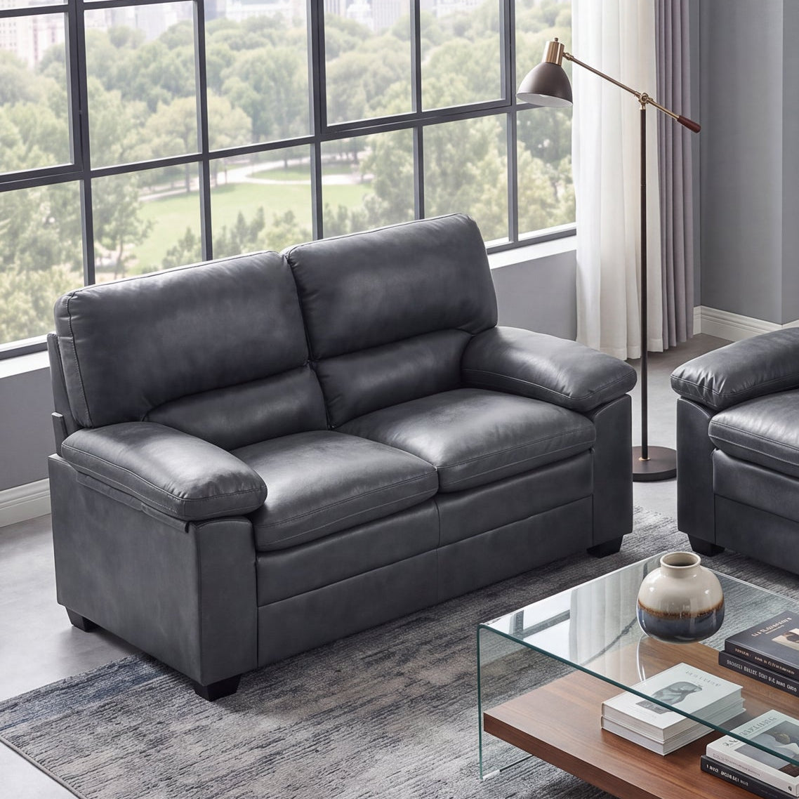A modern living room setup featuring a dark gray faux-leather loveseat and a matching three-seater sofa, arranged around a glass-top coffee table on a blue-gray rug. The room has gray walls, a large multi-pane window looking out onto green trees, and a small console table with framed art.